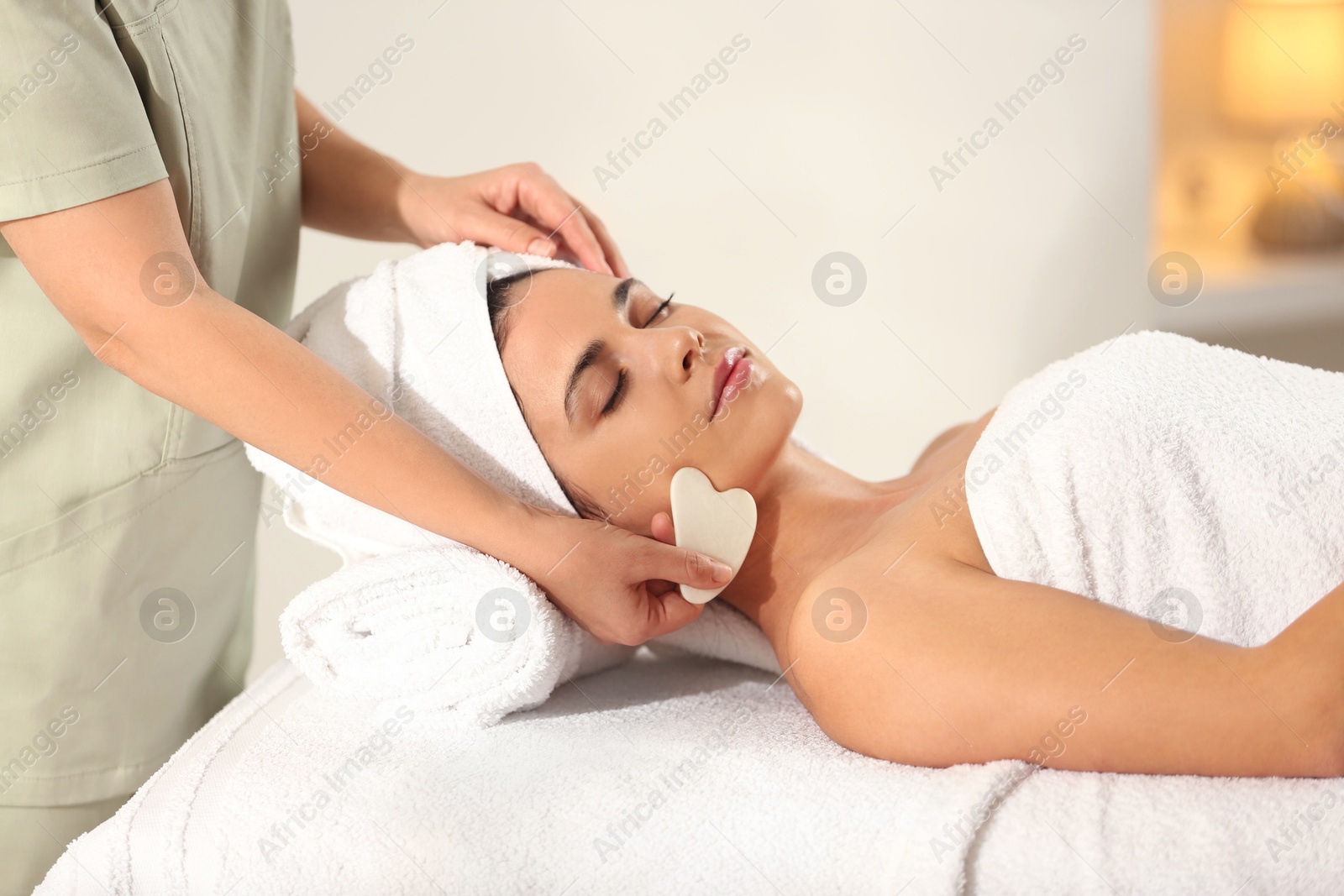 Photo of Young woman receiving facial massage with gua sha tool in spa salon