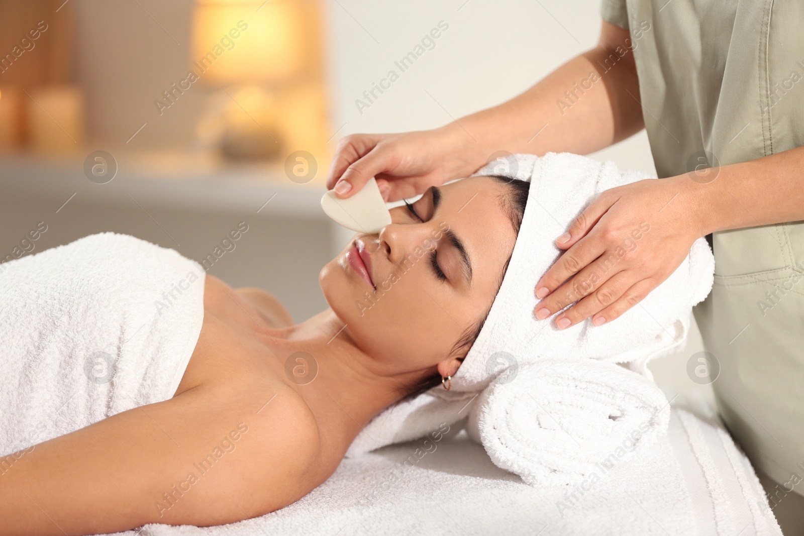 Photo of Young woman receiving facial massage with gua sha tool in spa salon