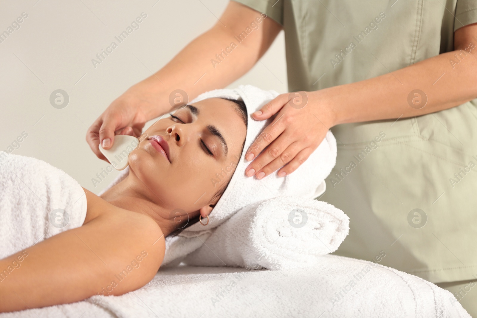 Photo of Young woman receiving facial massage with gua sha tool in spa salon