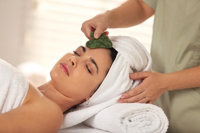 Photo of Young woman receiving facial massage with gua sha tool in spa salon