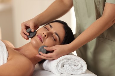 Photo of Young woman receiving facial massage with spa stones in salon