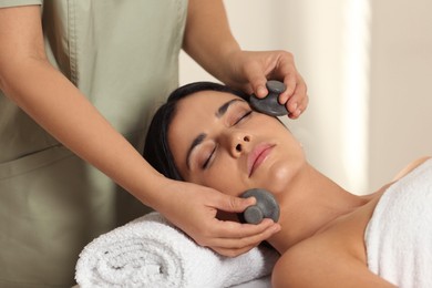 Photo of Young woman receiving facial massage with spa stones in salon