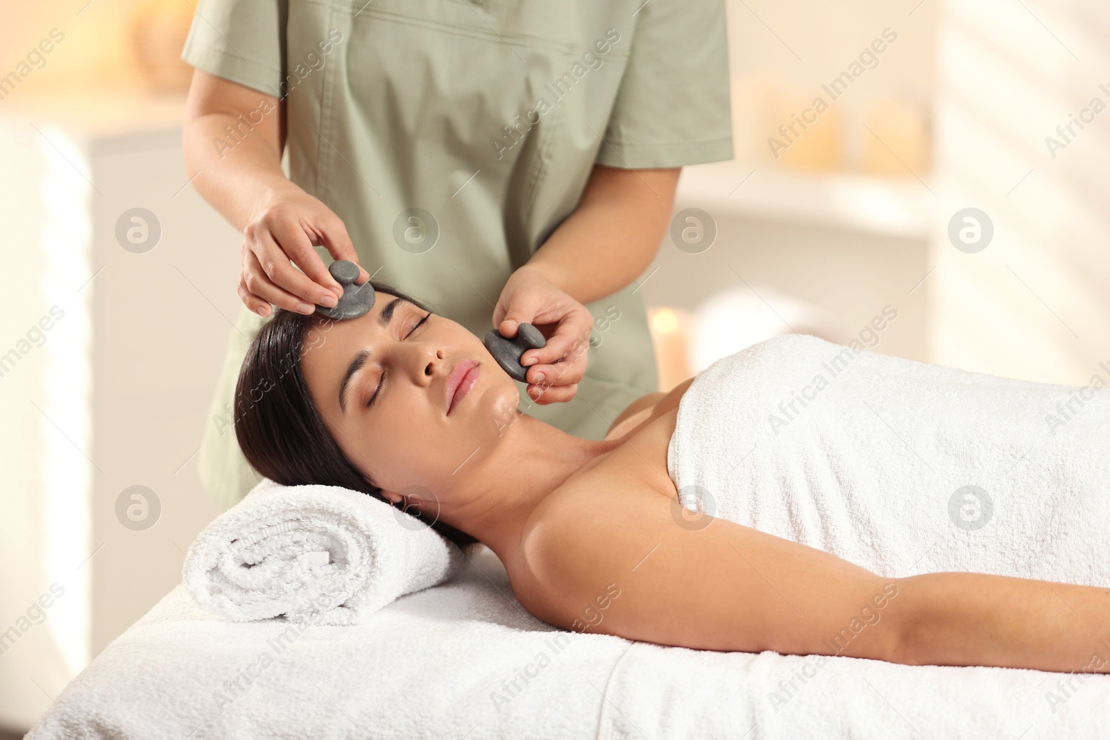 Photo of Young woman receiving facial massage with spa stones in salon