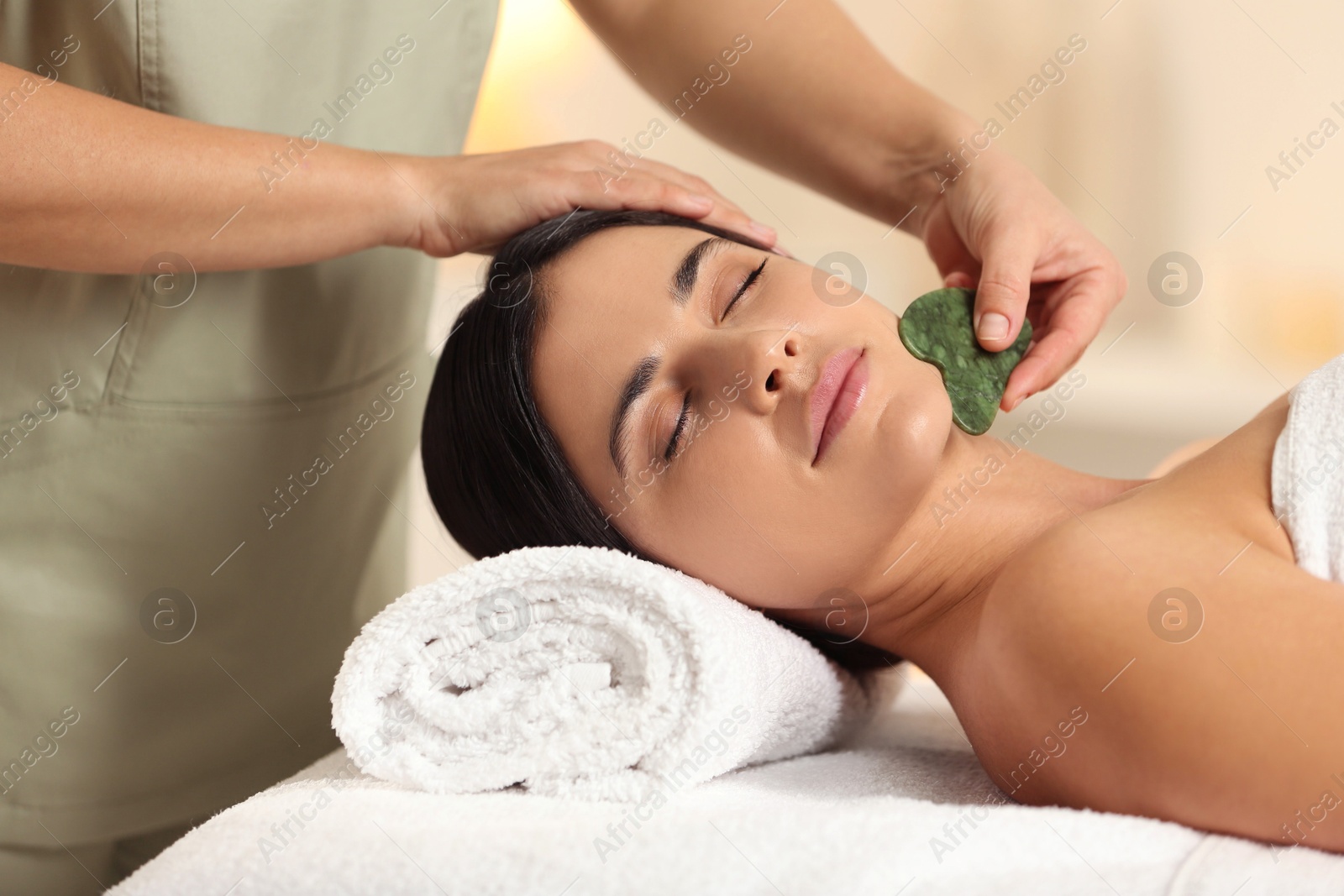 Photo of Young woman receiving facial massage with gua sha tool in spa salon