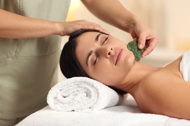 Photo of Young woman receiving facial massage with gua sha tool in spa salon