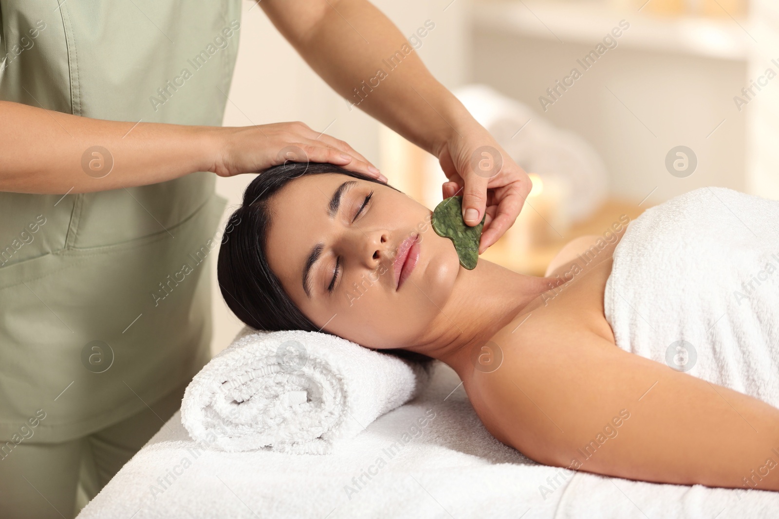 Photo of Young woman receiving facial massage with gua sha tool in spa salon