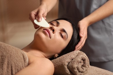 Photo of Young woman receiving facial massage with gua sha tool in spa salon