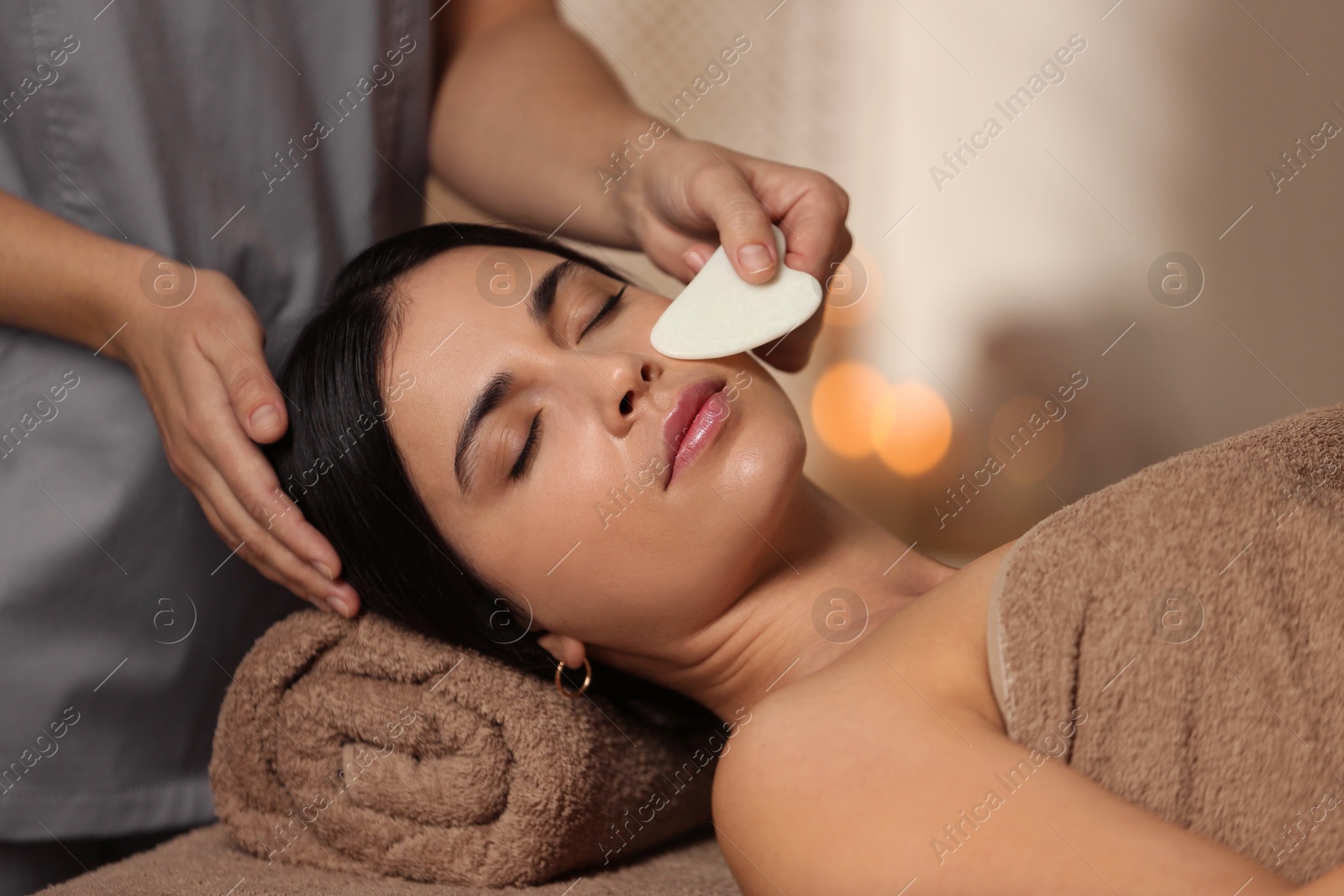 Photo of Young woman receiving facial massage with gua sha tool in spa salon