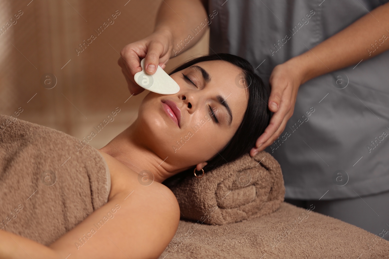 Photo of Young woman receiving facial massage with gua sha tool in spa salon