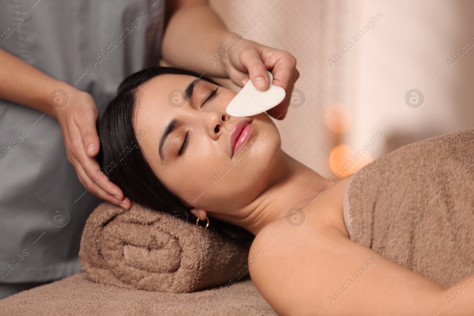 Photo of Young woman receiving facial massage with gua sha tool in spa salon