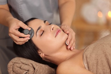 Photo of Young woman receiving facial massage with spa stone in salon