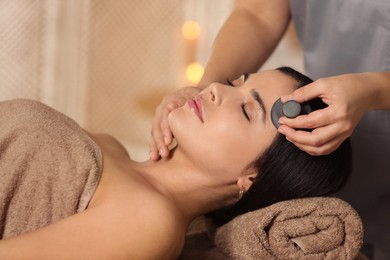 Photo of Young woman receiving facial massage with spa stone in salon