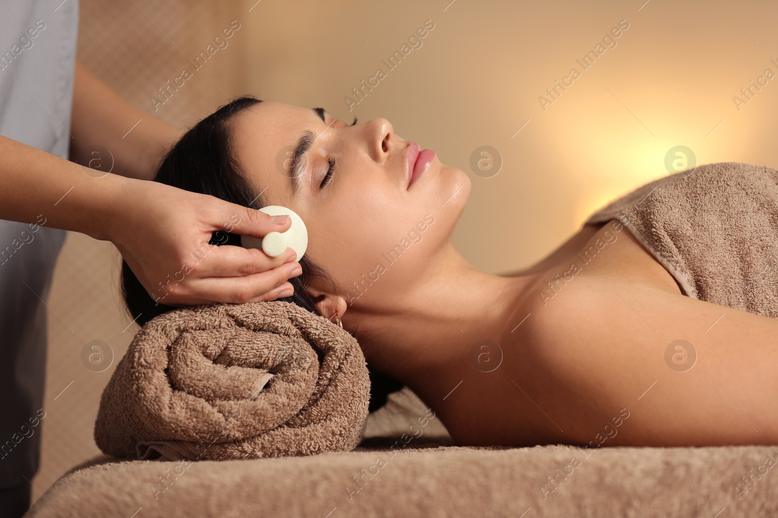 Photo of Young woman receiving facial massage with spa stone in salon