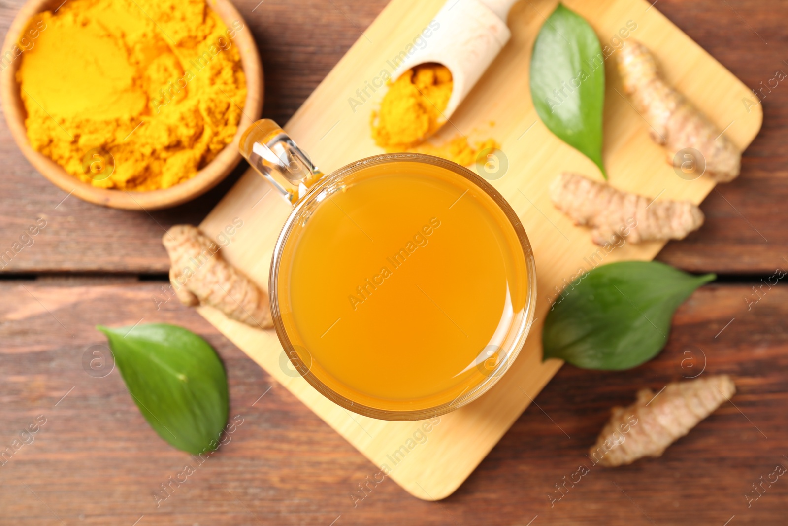 Photo of Aromatic turmeric tea in glass mug, roots, powder and green leaves on wooden table, flat lay