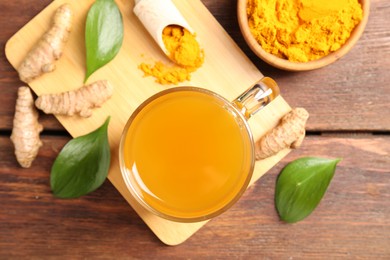 Photo of Aromatic turmeric tea in glass mug, roots, powder and green leaves on wooden table, flat lay