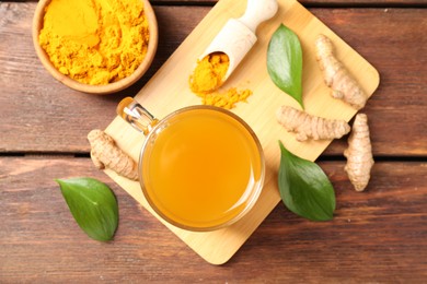 Photo of Aromatic turmeric tea in glass mug, roots, powder and green leaves on wooden table, flat lay