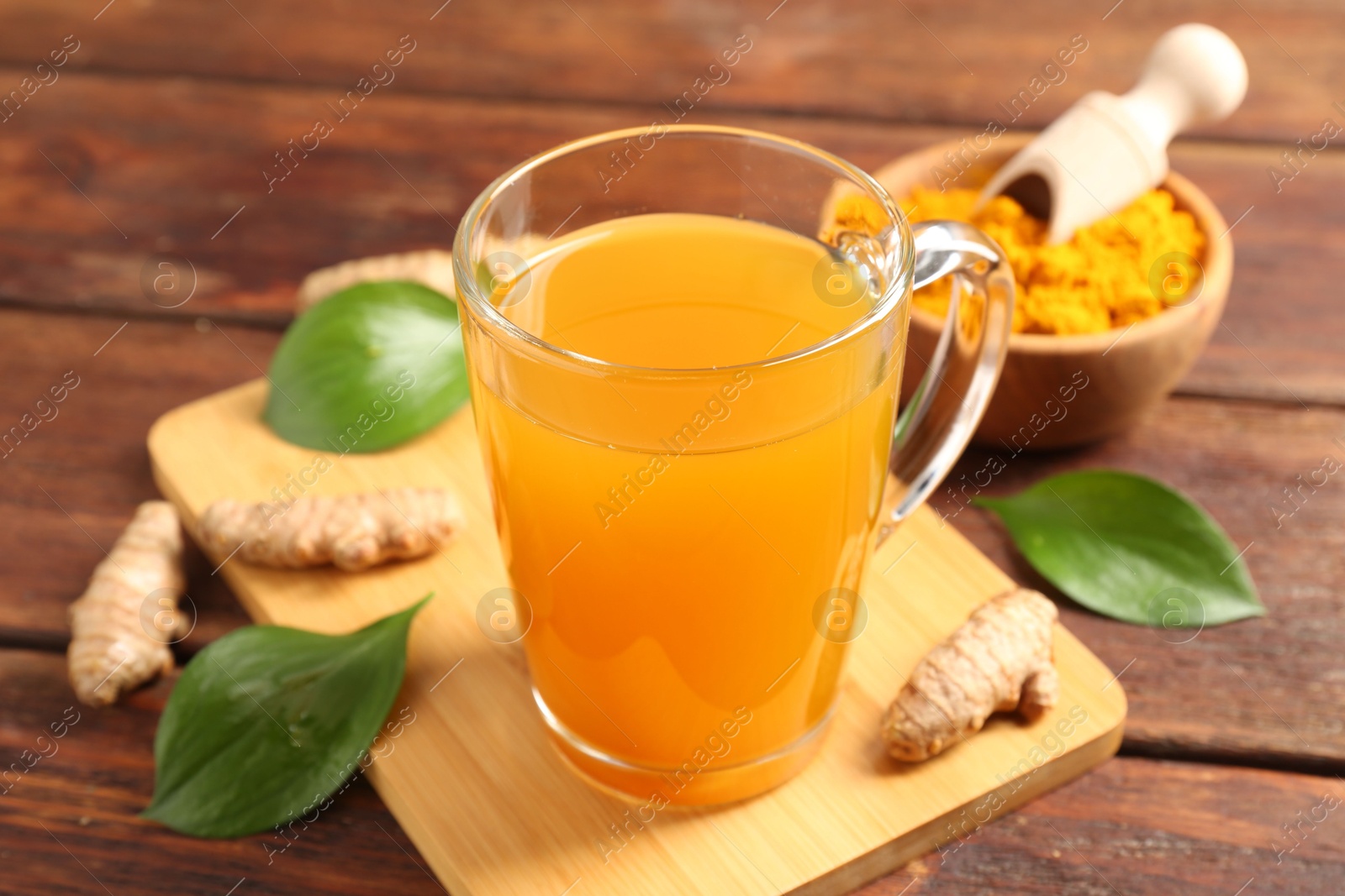 Photo of Aromatic turmeric tea in glass mug, roots, powder and green leaves on wooden table, closeup