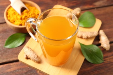 Photo of Aromatic turmeric tea in glass mug, roots, powder and green leaves on wooden table, closeup