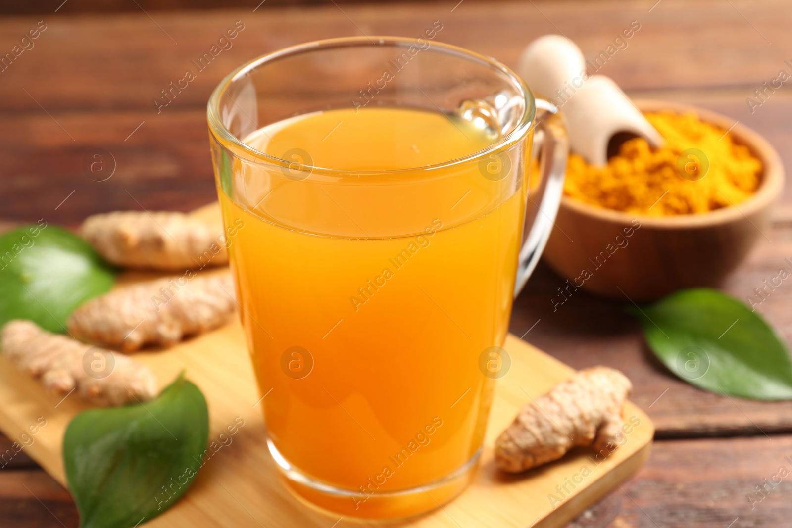 Photo of Aromatic turmeric tea in glass mug, roots, powder and green leaves on wooden table, closeup