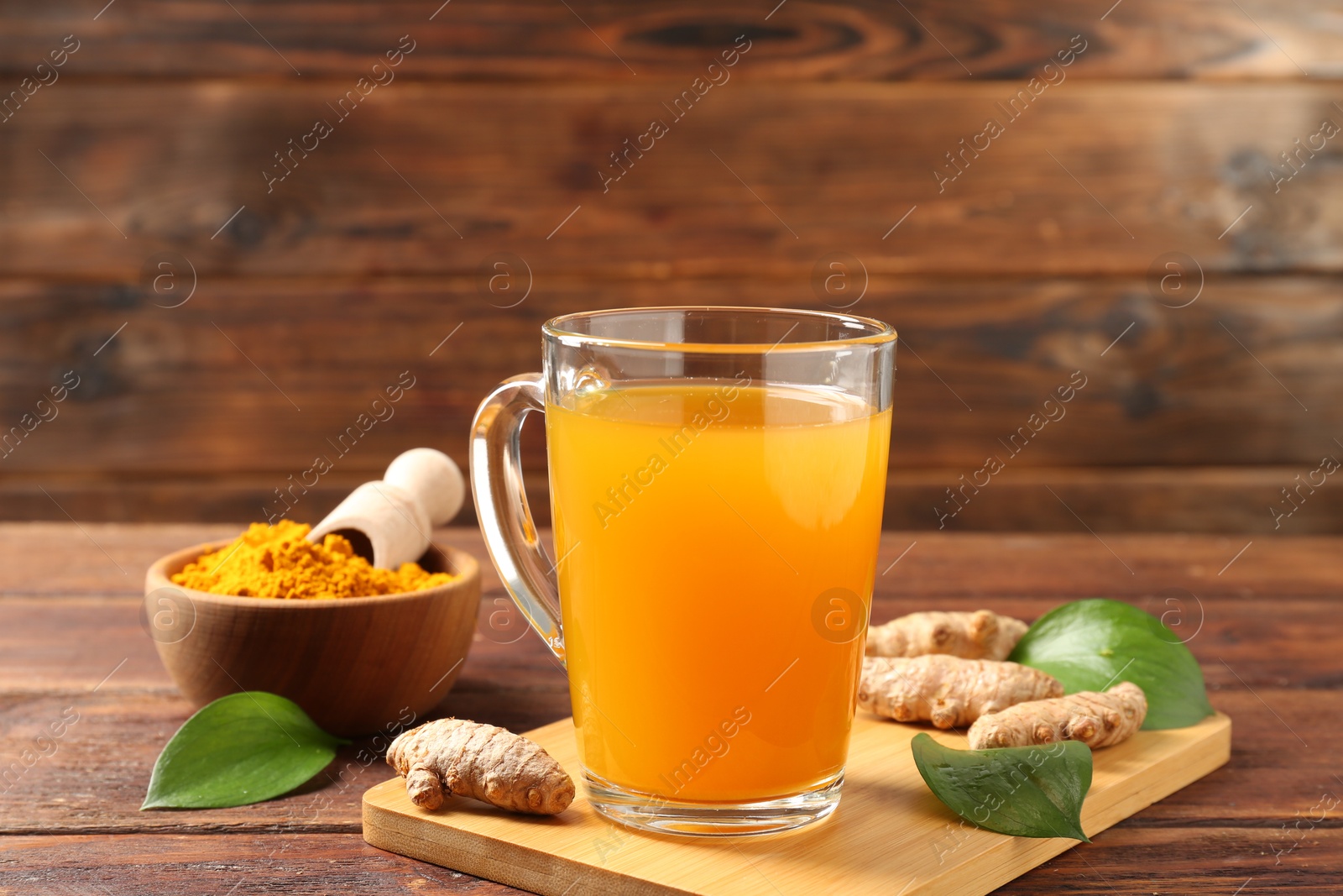 Photo of Aromatic turmeric tea in glass mug, roots, powder and green leaves on wooden table