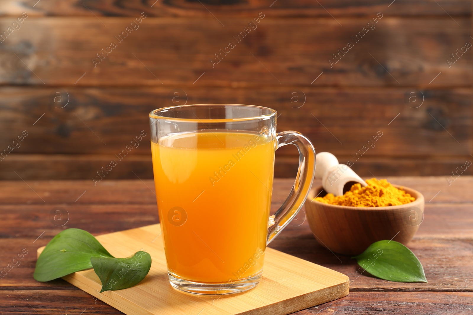 Photo of Aromatic turmeric tea in glass mug, roots, powder and green leaves on wooden table