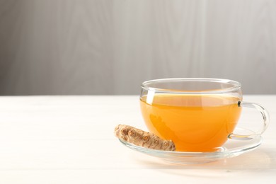 Photo of Aromatic turmeric tea in glass cup and root on white wooden table, closeup. Space for text