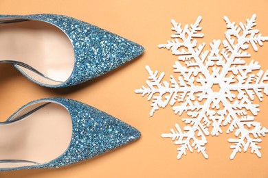 Shiny high heeled shoes and decorative snowflake on pale orange background, flat lay