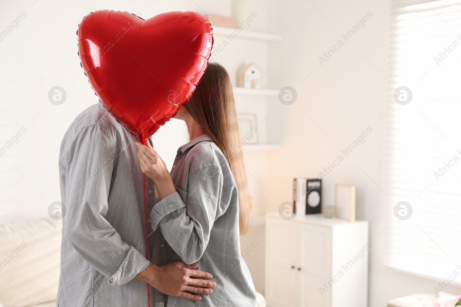 Photo of Lovely couple kissing behind heart shaped balloon indoors, space for text. Valentine's day celebration