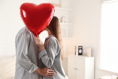 Photo of Lovely couple kissing behind heart shaped balloon indoors, space for text. Valentine's day celebration