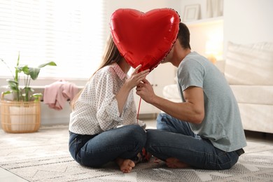 Photo of Lovely couple kissing behind heart shaped balloon indoors. Valentine's day celebration