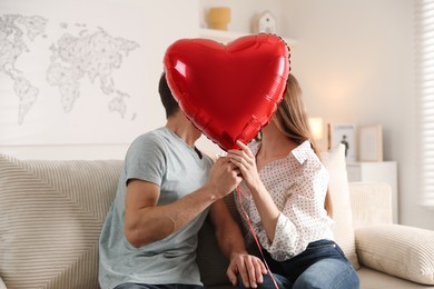 Photo of Lovely couple kissing behind heart shaped balloon on sofa indoors. Valentine's day celebration