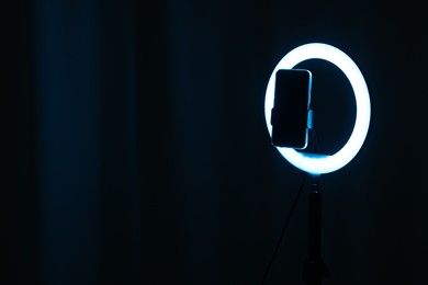 Photo of Glowing ring lamp with smartphone on black background. Space for text