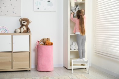 Photo of Little girl standing on step stool and reaching for toys on shelf indoors, back view