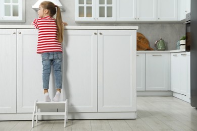 Photo of Little girl standing on step stool and reaching towards counter in kitchen, back view. Space for text