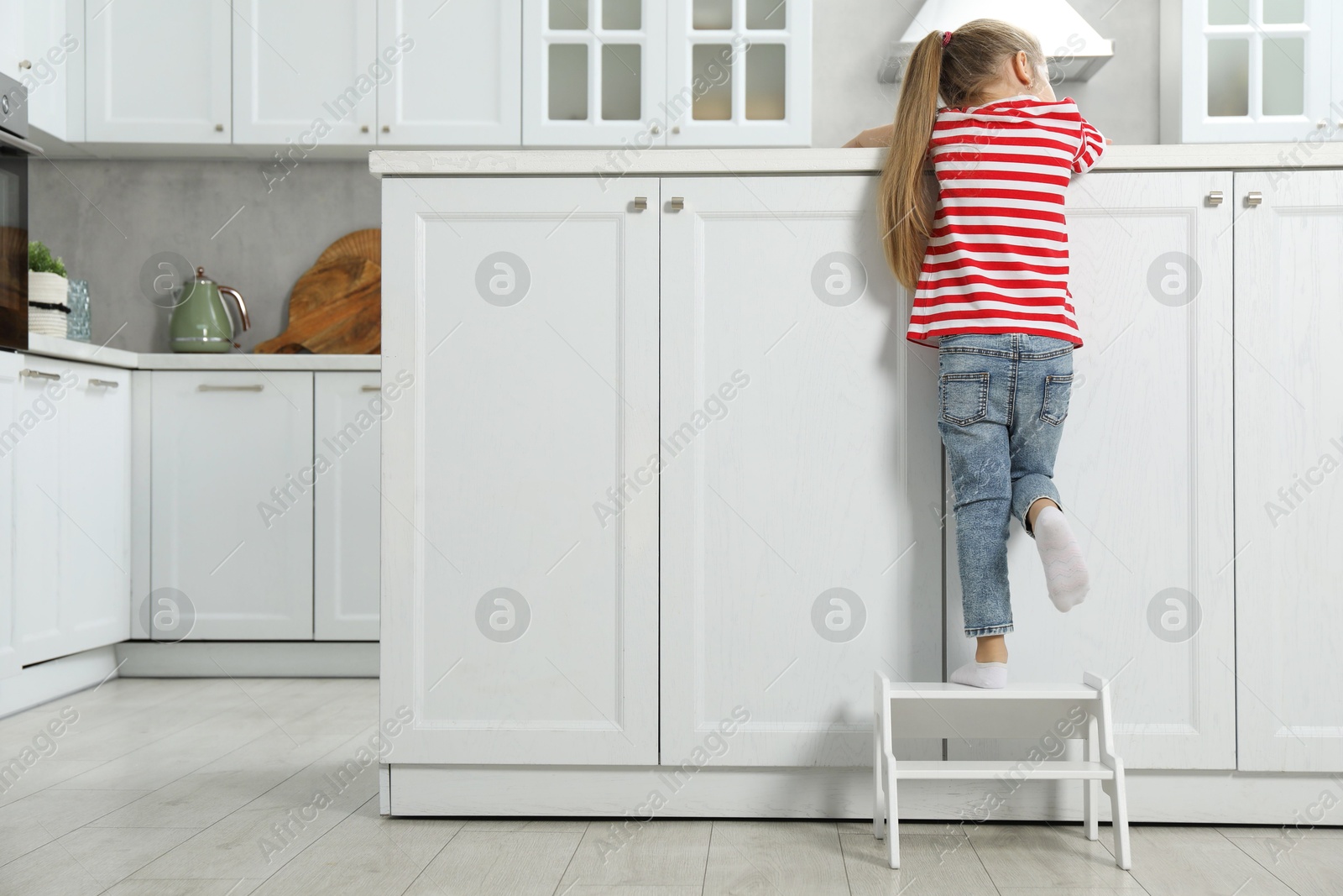 Photo of Little girl standing on step stool and reaching towards counter in kitchen, back view. Space for text