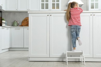 Photo of Little girl standing on step stool and reaching towards counter in kitchen, back view. Space for text