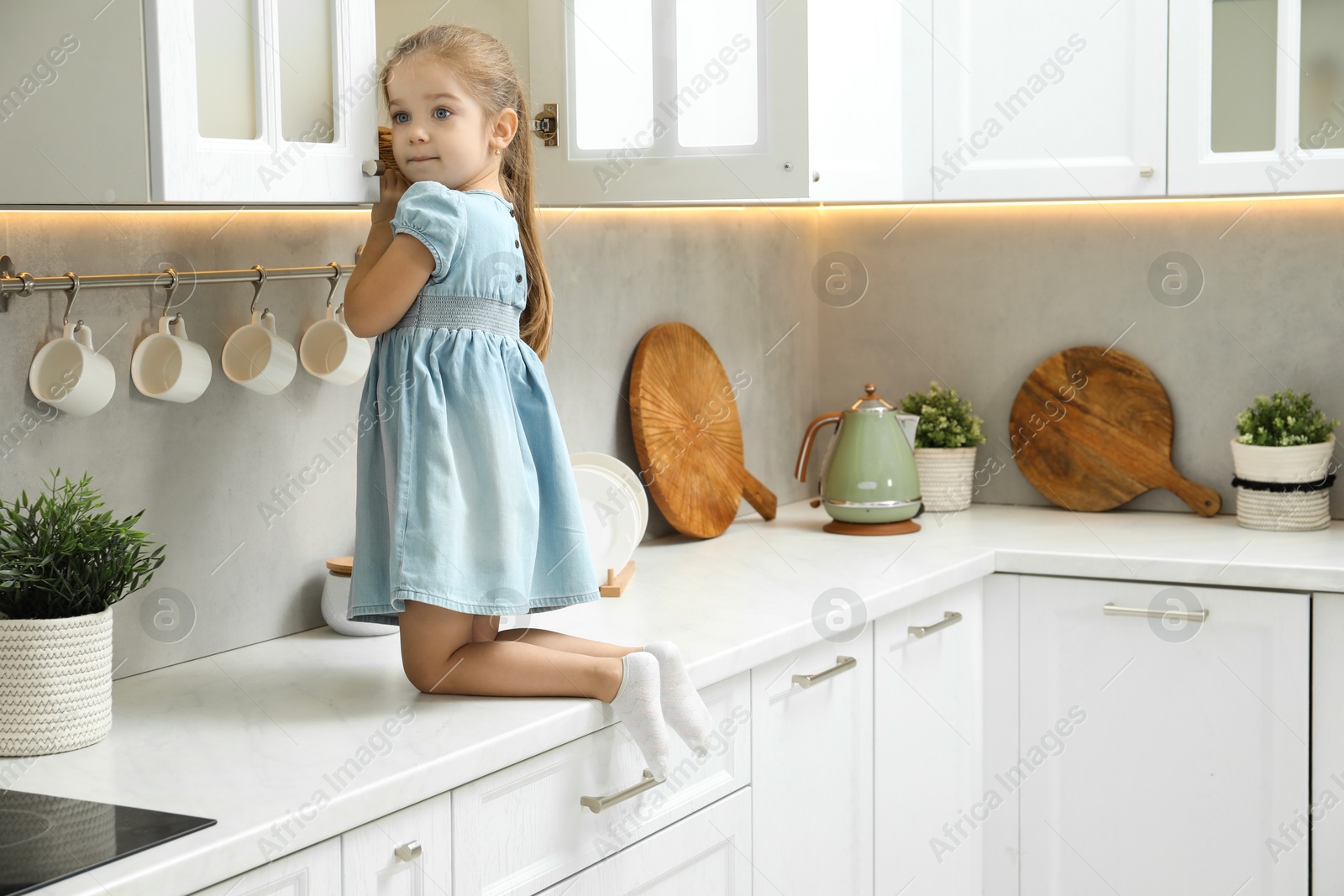 Photo of Little girl sitting on counter and reaching towards shelf in kitchen