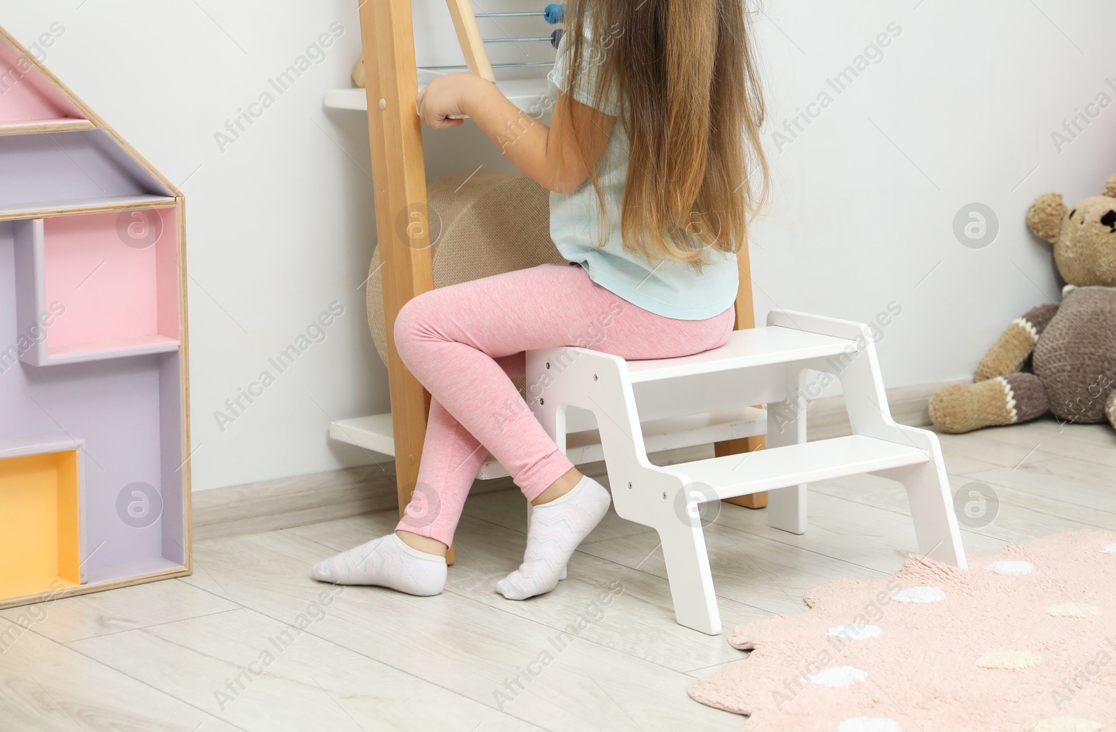 Photo of Little girl sitting on step stool near shelf with toys at home, back view