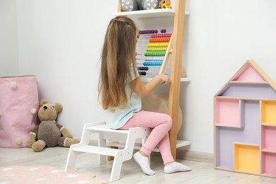 Photo of Little girl sitting on step stool near shelf with toys at home, back view