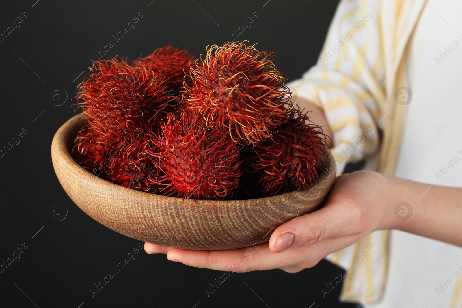 Photo of Woman holding fresh rambutans in bowl on black background, closeup