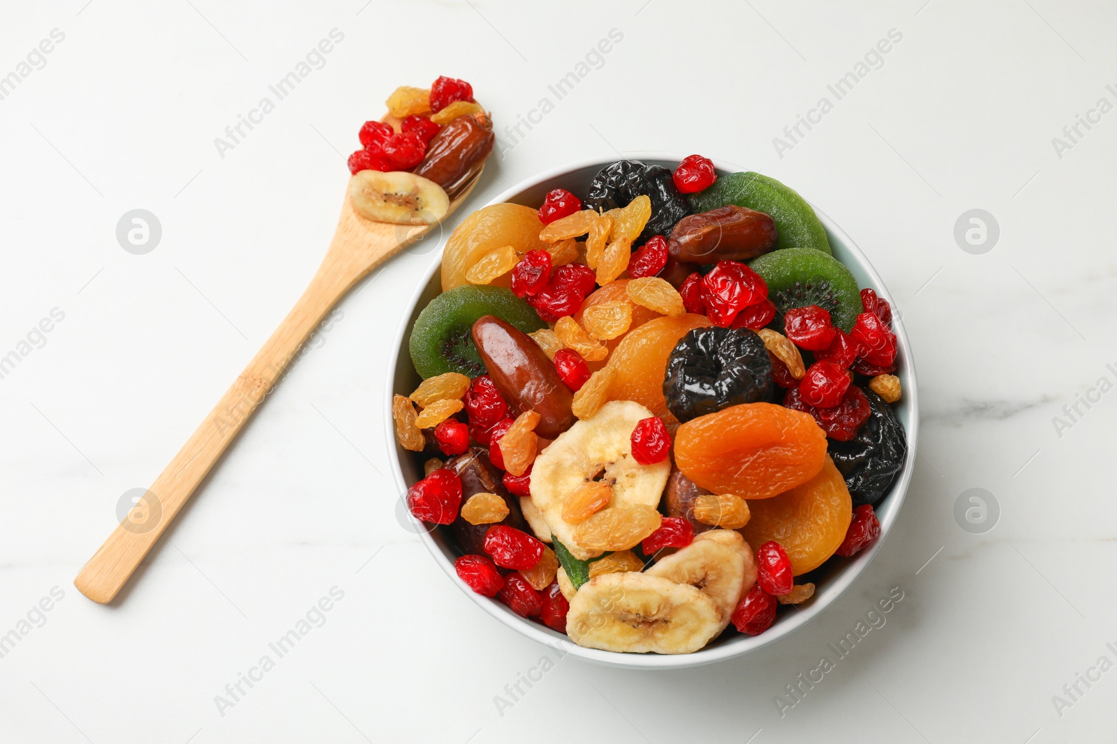 Photo of Mix of different dried fruits in bowl and spoon on white marble table, flat lay