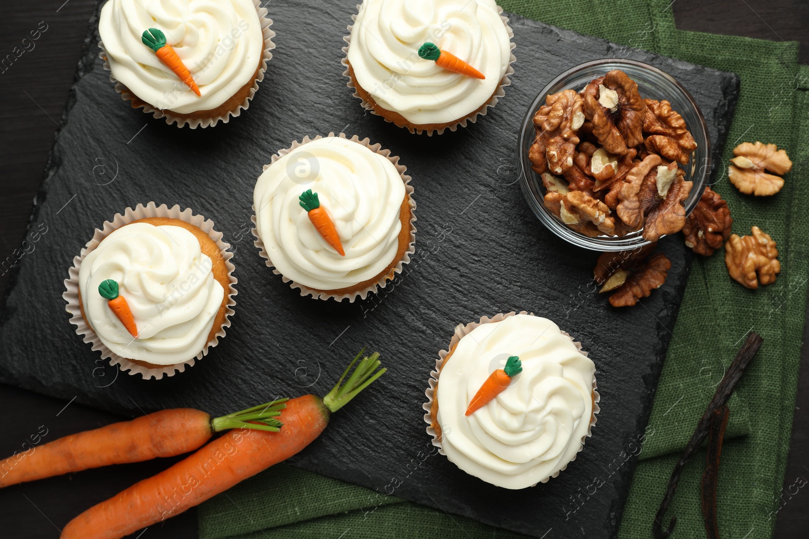 Photo of Tasty carrot muffins with cream, walnuts and fresh vegetables on black wooden table, flat lay