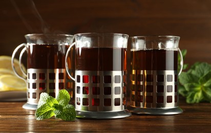 Photo of Glasses of hot tea in holders, mint and lemon on wooden table