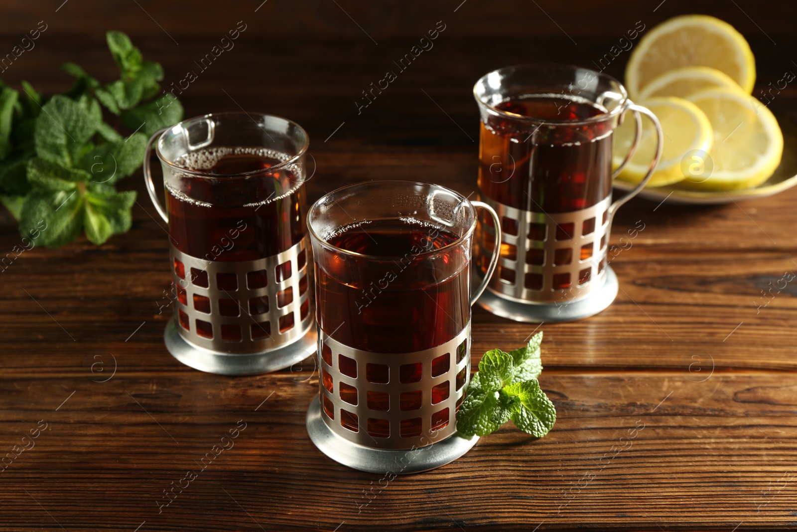 Photo of Glasses of hot tea in holders, mint and lemon on wooden table