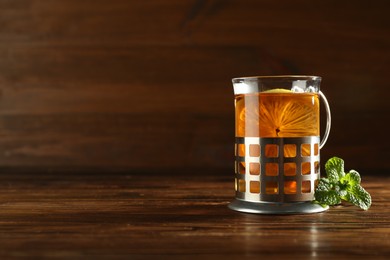 Photo of Glass of hot tea with lemon in holder and mint on wooden table, space for text