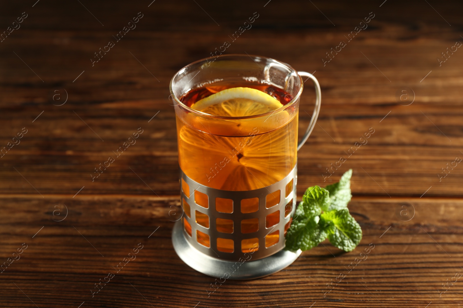 Photo of Glass of hot tea with lemon in holder and mint on wooden table, closeup