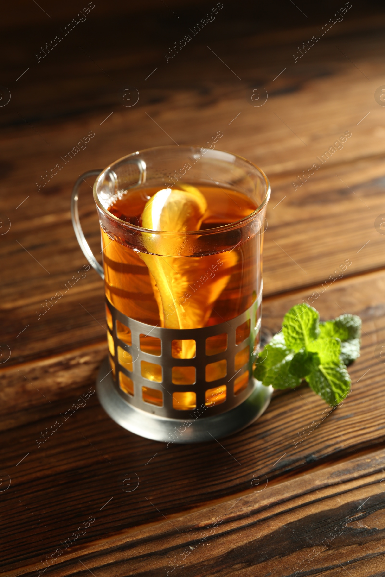 Photo of Glass of hot tea with lemon in holder and mint on wooden table