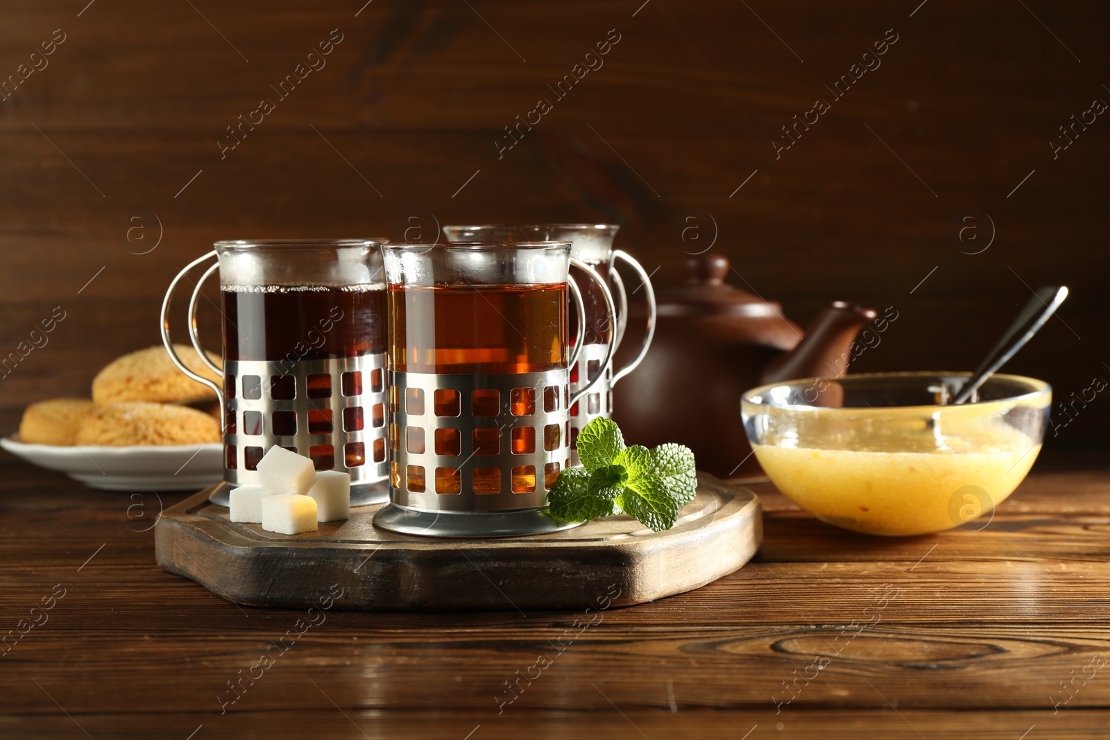 Photo of Glasses of hot tea in holders, sugar cubes, mint, honey and cookies on wooden table