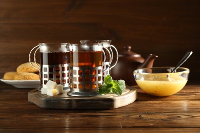 Photo of Glasses of hot tea in holders, sugar cubes, mint, honey and cookies on wooden table