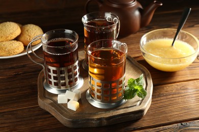 Photo of Glasses of hot tea in holders, sugar cubes, mint, honey and cookies on wooden table